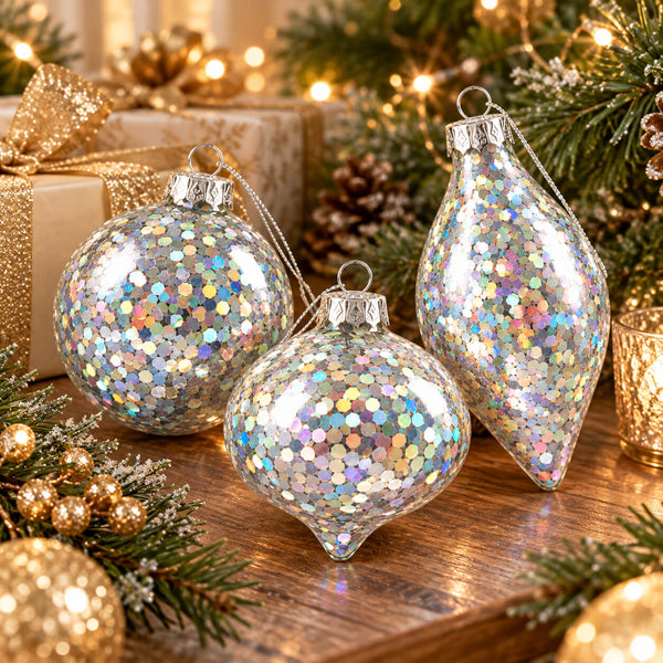 Three clear Christmas ornaments filled with iridescent holographic glitter, displayed on a wooden surface with festive greenery, gold ornaments, and wrapped gifts in the background.