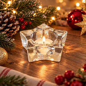 A clear glass star-shaped candle holder with a lit white tea light candle, placed on a wooden table with warm holiday lights, pine branches, red berries, and ornaments in the background.