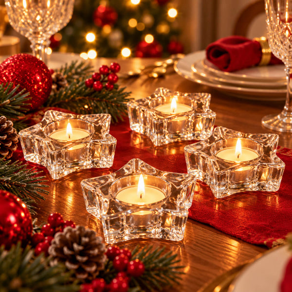 Four clear glass star-shaped candle holders with lit tealight candles arranged on a decorated Christmas table with red napkins, pinecones, greenery, and warm blurred lights in the background.