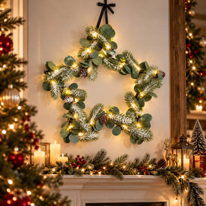 A star-shaped Christmas wreath with green eucalyptus branches, white faux snow, warm white string lights, and small pinecones, hanging on a wall above a decorated fireplace mantel.