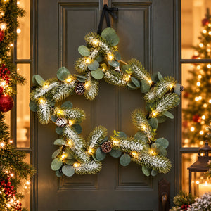 A festive star-shaped holiday wreath hanging on a dark gray front door. The wreath is made of green eucalyptus branches with white faux snow, warm white string lights, and small pinecones.