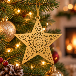 A gold metal star-shaped Christmas ornament with intricate cutout patterns hangs on a decorated Christmas tree in front of a warm, blurred fireplace background.