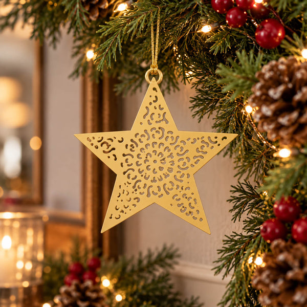 A gold metal star-shaped Christmas ornament with intricate cutout patterns hangs from a decorated Christmas tree branch with pinecones, red berries, and warm string lights.