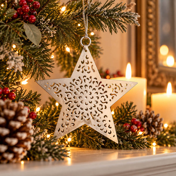 A silver metal star-shaped Christmas ornament with intricate cutout patterns hangs on a decorated Christmas tree branch with pinecones, red berries, and warm string lights.