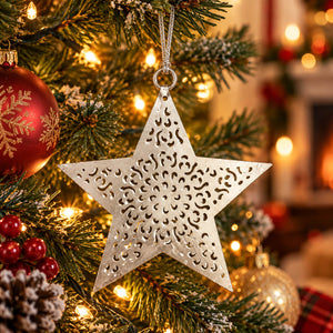 A silver metal star Christmas ornament with intricate cutout patterns hangs on a decorated Christmas tree with warm lights and holiday decorations in the background.