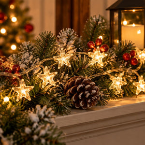 Warm white star-shaped string lights woven through a green pine garland with faux snow, red berries, and a pinecone, displayed on a mantel in a cozy holiday setting.