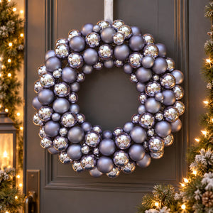 A festive Christmas wreath decorated with a mix of matte lavender-gray and reflective silver ornaments, hanging on a dark gray paneled door with warm holiday lights visible in the background.