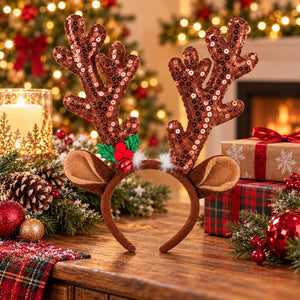 A festive Christmas headband with sequined brown reindeer antlers, plush tan ears, and a small red holly accent, displayed on a wooden table in a decorated holiday setting with a blurred Christmas tree and wrapped gifts in the background.