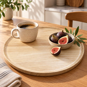 A round light wood serving tray on a wooden table, styled with a beige ceramic coffee mug, a small bowl of figs, and a green leafy sprig in a bright indoor setting.