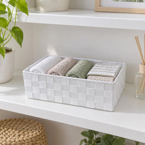 A white woven rectangular storage basket holding five folded towels in neutral and sage green tones, styled on a white shelf with a plant and diffuser nearby.