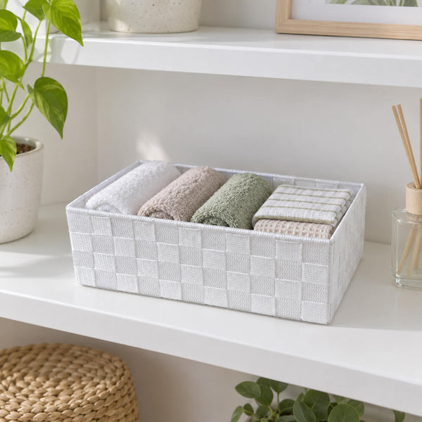 A white woven rectangular storage basket holding five folded towels in neutral and sage green tones, styled on a white shelf with a plant and diffuser nearby.