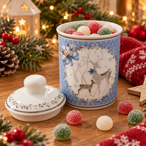A festive ceramic candy jar with a matching lid, decorated with blue and white holiday motifs of reindeer and snowflakes, filled with colorful frosted candies on a wooden table surrounded by Christmas decorations.