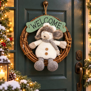 A festive holiday wreath hanging on a dark green front door, featuring a plush winter animal character in a cream coat and gray scarf, with a green 'WELCOME' sign above it. The scene is decorated with warm lights, pine branches, and snow-dusted foliage.