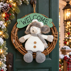 A plush snowman decoration hanging on a rustic grapevine wreath with a green 'WELCOME' sign, displayed on a dark green front door decorated with holiday lights and garlands.