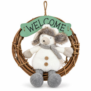A plush winter snowman hanging from a woven wicker wreath with a green 'WELCOME' sign. The snowman has white fur, gray felted legs and scarf, and brown buttons.