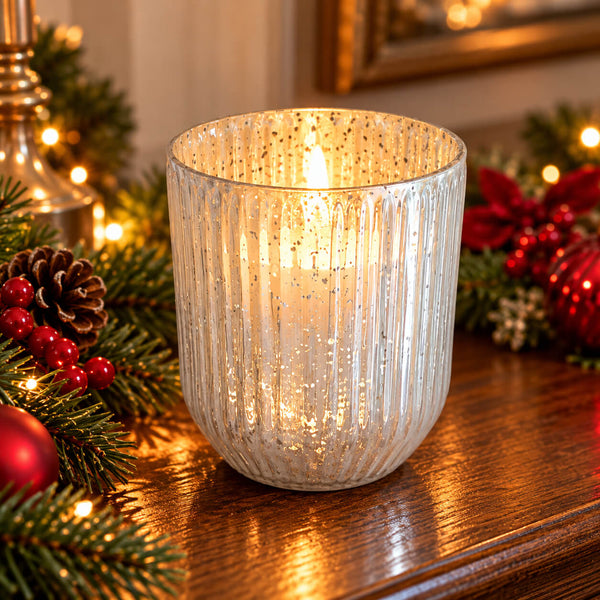 A ribbed glass candle holder with a warm white glitter finish, lit with a candle inside, set on a wooden table surrounded by Christmas decorations including pine branches, red berries, and ornaments.