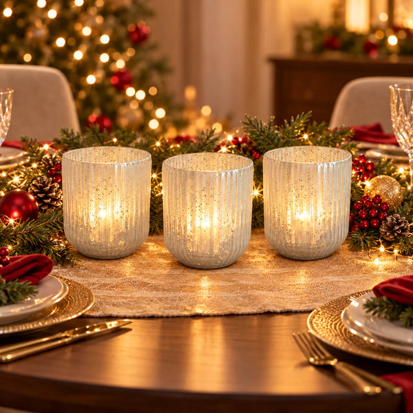 Three ribbed glass candle holders with warm white LED lights, styled on a wooden dining table with a Christmas garland, pinecones, red ornaments, and gold cutlery.