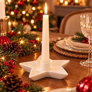 A white ceramic star-shaped candle holder with a lit white taper candle, set on a wooden dining table decorated for Christmas with pine branches, red berries, and warm string lights.