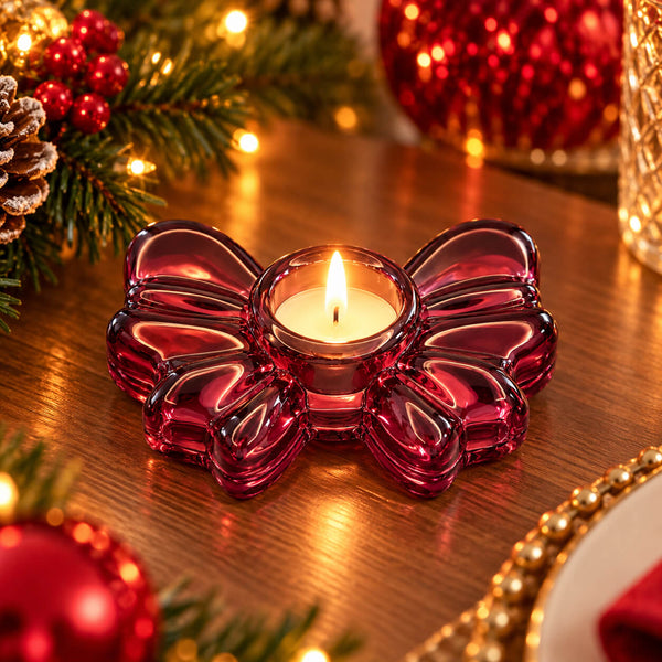 A small lit tealight candle in a translucent dark red bow-shaped glass holder, placed on a wooden table with festive Christmas decorations in the background.