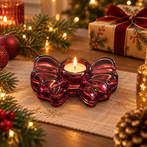 A translucent burgundy glass bow-shaped candle holder with a lit tealight candle, styled on a burlap runner with Christmas lights, pine branches, ornaments, and a wrapped gift box in the background.
