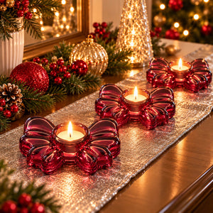 Three red glass bow-shaped candle holders with lit tealight candles arranged on a silver glitter table runner in a festive holiday setting with pine garlands and ornaments.