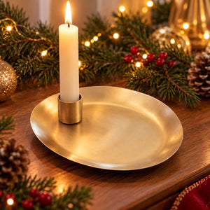 A round gold-toned metal tray holding a lit white candle in a matching candle holder, styled on a wooden surface with Christmas greenery, pinecones, red berries, and warm string lights in the background.