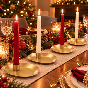 Four gold candlesticks with red and white taper candles arranged on a decorated Christmas table runner, with warm holiday lights and a blurred festive background.