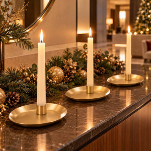 Three gold candle holders with lit white taper candles arranged on a dark marble countertop with holiday greenery and gold ornaments in a warmly lit interior setting.