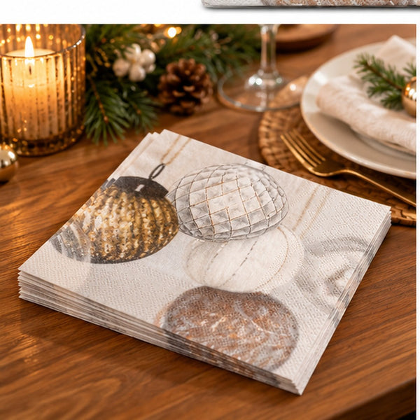 A stack of printed paper napkins on a wooden table, featuring a holiday design with metallic gold and silver ornaments. The background shows a cozy, decorated dining setting with a lit candle, pine branches, and plates.