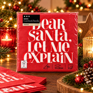A package of red paper napkins wrapped in clear plastic, standing on a wooden table with holiday decorations. The napkin package reads 'Dear Santa, Let Me Explain' in large white text.