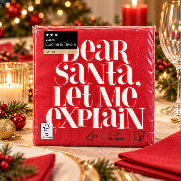 A pack of red paper napkins wrapped in clear plastic, featuring the text 'Dear Santa, Let Me Explain' in large white letters. The napkin pack is displayed on a decorated Christmas table with candles, pine branches, and red napkins.