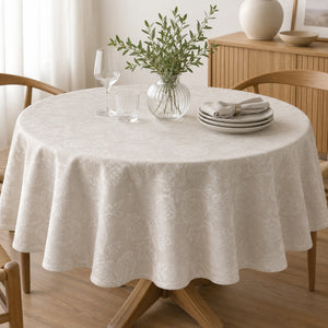 A round dining table covered with a light beige tablecloth featuring a subtle tonal floral pattern. The table is styled with a glass vase of greenery, clear drinking glasses, and a stack of gray plates with a folded napkin, set in a warm, softly lit dining room with wooden chairs and a cabinet in the background.