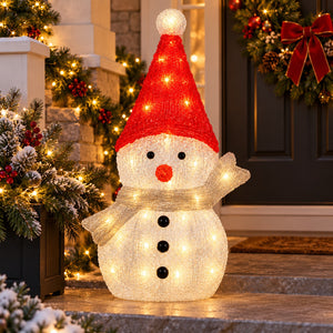 A large illuminated snowman decoration standing on a front porch, wearing a red hat with a white pom-pom and a glowing cream body and scarf. The scene is decorated with holiday garlands, lights, and a wreath in the background