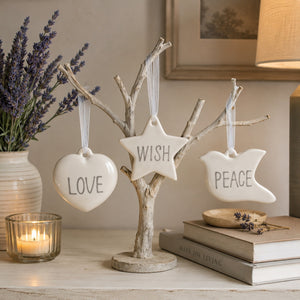 Three white ceramic hanging ornaments shaped like a heart, star, and dove, each printed with a word (LOVE, WISH, PEACE), displayed on a small rustic tree stand on a tabletop with candles, books, and lavender in a softly lit room.