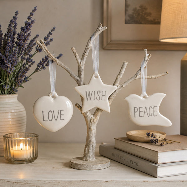 Three white ceramic hanging ornaments shaped like a heart, star, and dove, each printed with a word (LOVE, WISH, PEACE), displayed on a small rustic tree stand on a tabletop with candles, books, and lavender in a softly lit room.