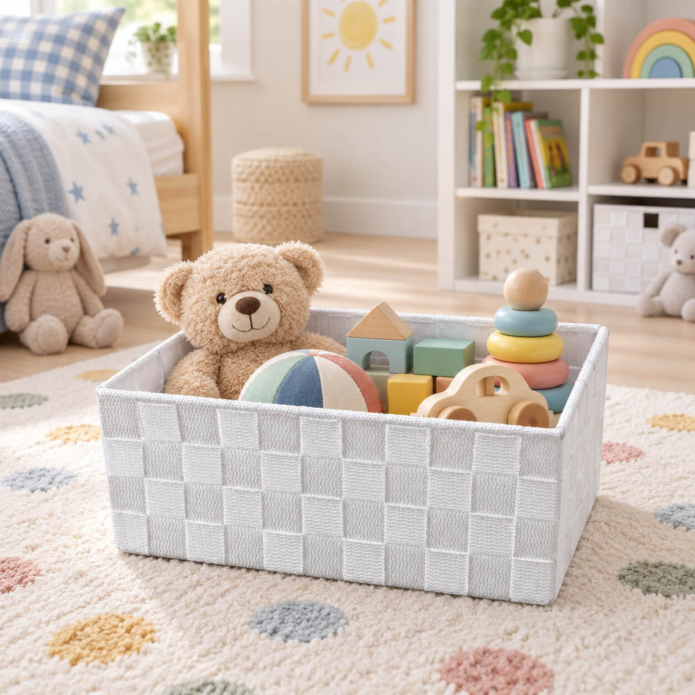 A white woven rectangular storage basket holding five folded towels in neutral and sage green tones, styled on a white shelf with a plant and diffuser nearby.