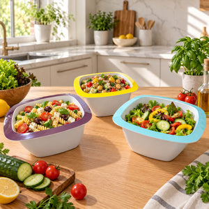 Three white square salad bowls with colorful rims are filled with fresh pasta salad and green salad on a wooden kitchen table, surrounded by vegetables and kitchen props.