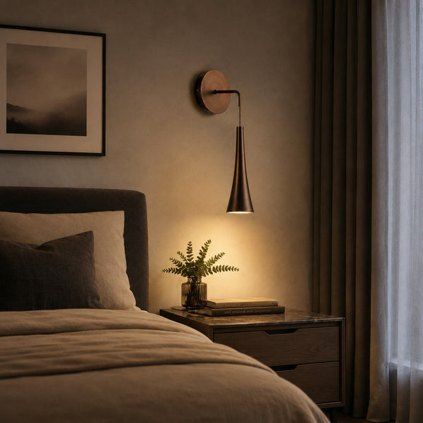 A modern wall-mounted sconce with a tapered metal shade and circular backplate, casting a warm light next to a bed in a styled bedroom setting.