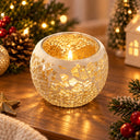 A round mosaic crackle glass candle holder with a lit tealight candle, set on a wooden table surrounded by festive Christmas decorations including pine branches, lights, and a small house ornament.