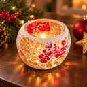 A round mosaic candle holder with a crackled white, gold, and red pattern, lit with a warm candle flame on a wooden table with festive Christmas decorations in the background.