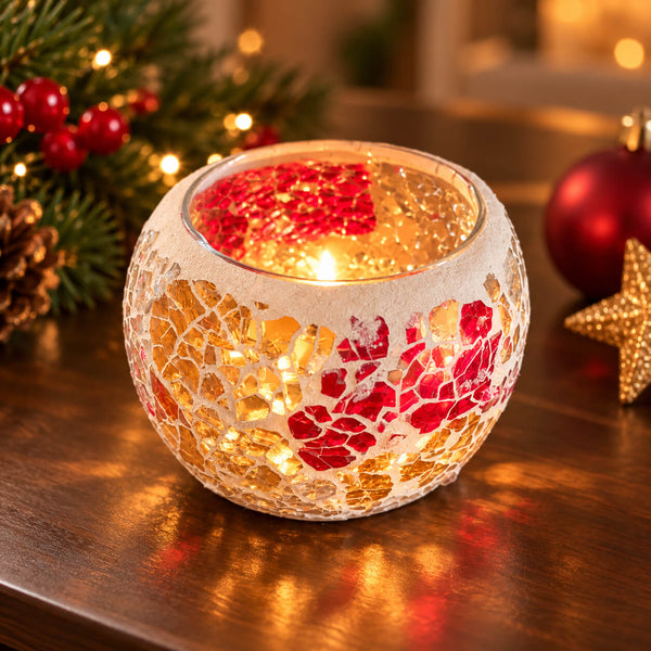 A round mosaic candle holder with a crackled white, gold, and red pattern, lit with a warm candle flame on a wooden table with festive Christmas decorations in the background.