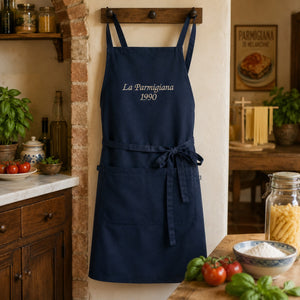 Navy blue bib apron with off-white “La Parmigiana 1990” embroidery, adjustable straps, waist ties, and a large front pocket on a white background.
