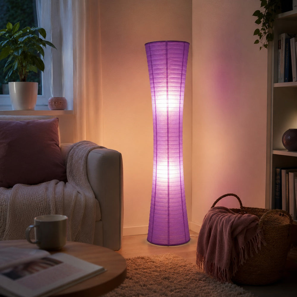 A tall, slender floor lamp with a purple textured shade stands in a cozy living room corner, casting a warm glow. The room includes a beige armchair with a blanket, a coffee table with a mug, a woven basket, and a bookshelf in the background.