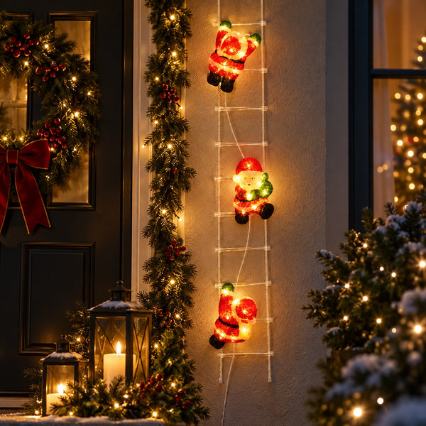 Three illuminated Christmas Santa Claus figures climbing a white ladder decoration on a house exterior at night, surrounded by holiday garlands, a wreath, lanterns, and a lit Christmas tree.