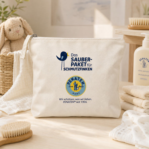 A cream-colored canvas pouch with printed branding for 'Das Sauber-Paket für Schmutzfinken' and 'PENATEN BABY' sits on a light wooden table surrounded by baby-themed props like a plush bunny, folded towels, and a bottle, creating a warm, soft-focus lifestyle setting.