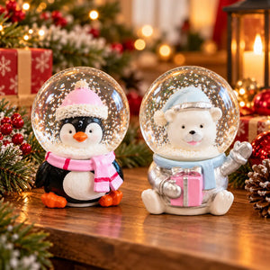 Two Christmas snow globes on a wooden table, one featuring a penguin in a pink winter hat and scarf, and the other featuring a white polar bear in a blue winter hat holding a pink gift box. The background is decorated with pine branches, red ornaments, and a warm lit lantern.