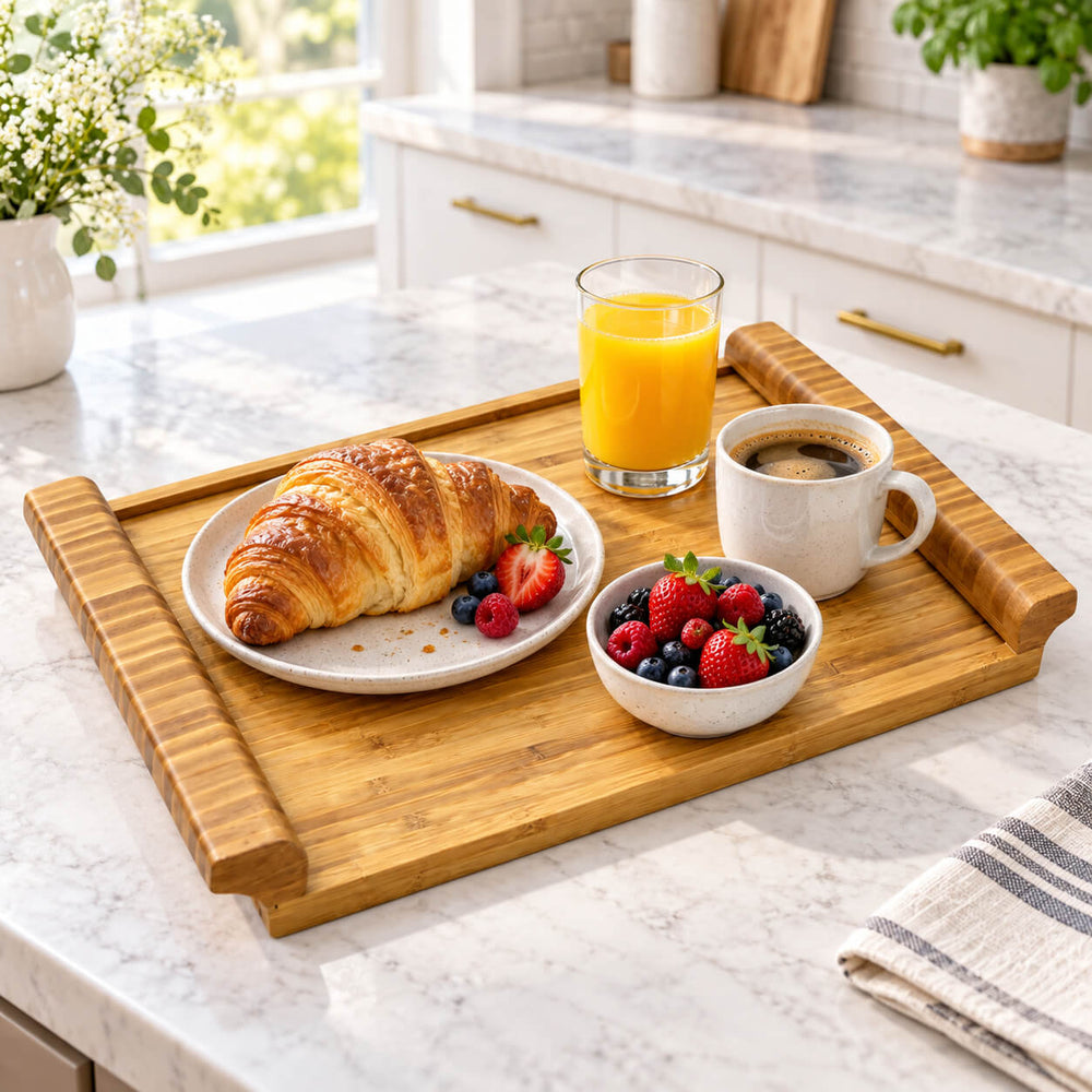 A rectangular wooden serving tray with raised edges sits on a white marble kitchen counter, styled with a croissant on a small plate, a glass of orange juice, a cup of coffee, and a bowl of fresh berries.