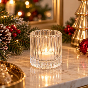 A clear ribbed glass candle holder with a lit white candle inside, placed on a marble surface with Christmas decorations in the background.