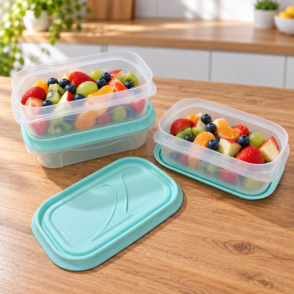 Three clear plastic rectangular food storage containers with light blue silicone lids are arranged on a wooden countertop, with one container open and filled with assorted fruit. A matching lid sits in the foreground.