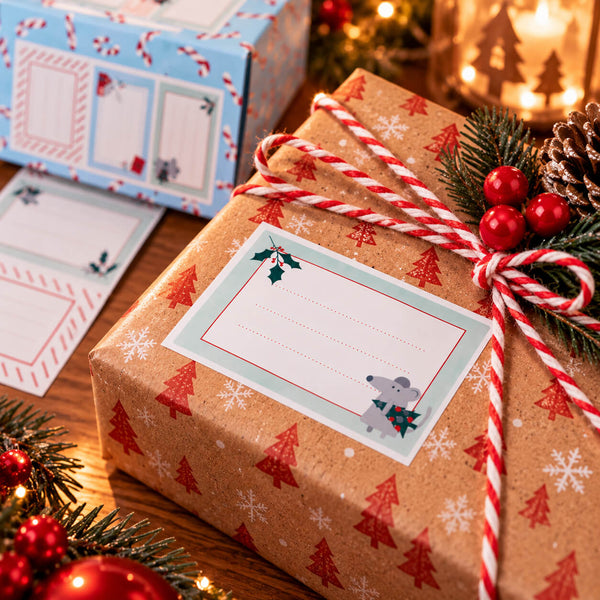 A wrapped Christmas gift box in tan kraft paper with a red and white holiday tree print, tied with a red-and-white striped ribbon and decorated with pine branches, red berries, and a gift tag. A matching gift box and gift tag are visible in the background on a wooden surface with warm holiday lights.